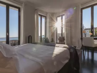 Una camera da letto soleggiata con molte grandi finestre. Al centro della stanza si trova un grande letto rivestito di bianco con vista sul mare.  