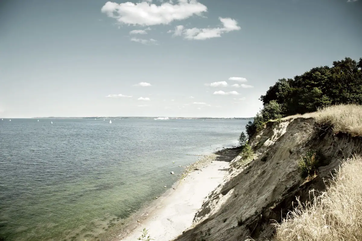 Strand mit Wasser und sandiger Klippe im Vordergrund.