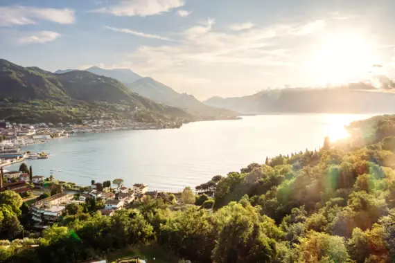 L'immagine mostra una suggestiva veduta del Lago di Garda, circondato da una vivace cittadina e da montagne verdeggianti sullo sfondo. Il sole del mattino illumina la scena, mentre una luce soffusa illumina la superficie dell'acqua e avvolge i paesaggi e gli edifici circostanti in un bagliore dorato. Questa atmosfera pacifica è esaltata dalla superficie calma dell'acqua e dalla vegetazione lussureggiante, che insieme creano un'atmosfera di relax e vicinanza alla natura.