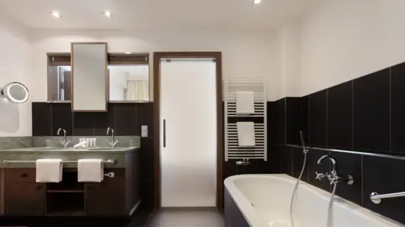 Un bagno con piastrelle scure, una vasca da bagno e un lavabo con due lavabi. Sopra la vasca è appeso uno scaldasalviette con due asciugamani bianchi.