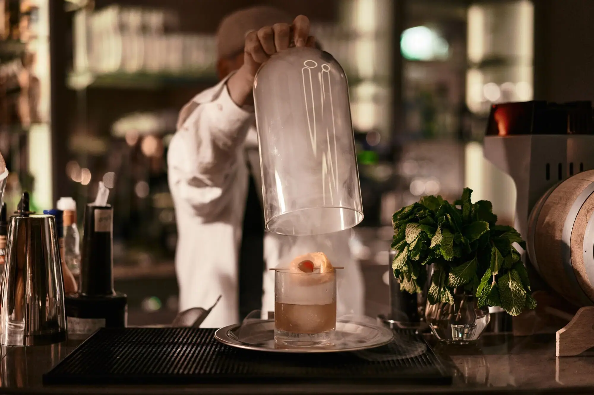 Person nimmt eine Glashaube über einem dampfenden Glas mit einem Drink hoch, umgeben von einer Bar mit einer Pflanze auf dem Tresen in einem Innenraum.