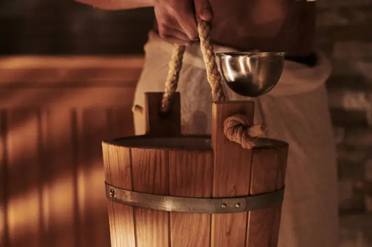 Primo piano di una mano che tiene un secchio da sauna in legno, dotato di un mestolo in metallo. Il secchio è tenuto da una corda robusta, che crea un'autentica atmosfera da sauna. 