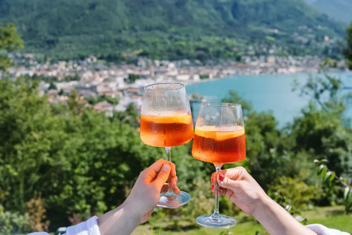 Aperol Spritz Due mani reggono dei calici da vino contenenti un liquido arancione, sullo sfondo di un lago.