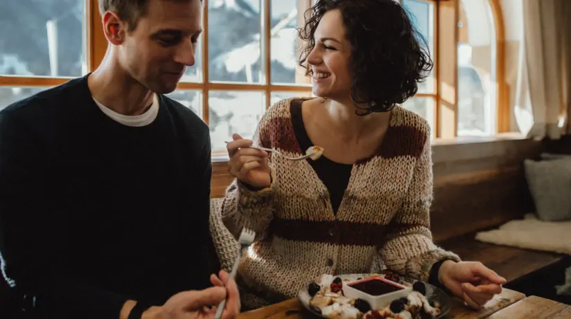 Un uomo e una donna seduti a un tavolo che mangiano cibo, con una torta di compleanno visibile.