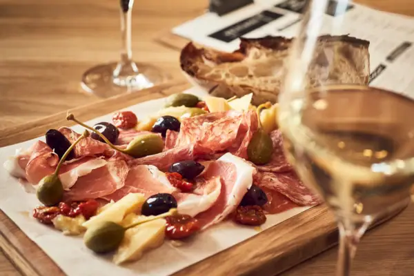 Ein Holzbrett mit Schinken und Oliven und einem Glas Weißwein im Vordergrund..