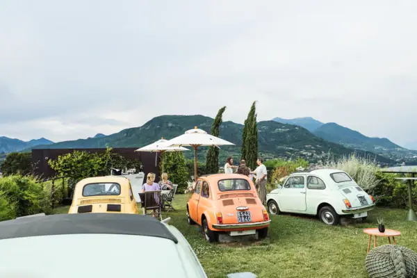 Eine Gruppe von Fiat 500 Autos auf dem Rasen geparkt, umgeben von Menschen, die sitzen und stehen. Sie halten jeweils Getränke in der Hand. Sonnenschirme sind aufgestellt. Im Hintergrund sieht man die italienischen Berge.
