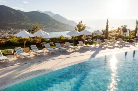 Piscina con sedie e ombrelloni in riva al mare, circondata da montagne e alberi.