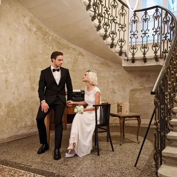 Un uomo in abito e una donna in abito da sposa seduti su una sedia in una stanza con scale.