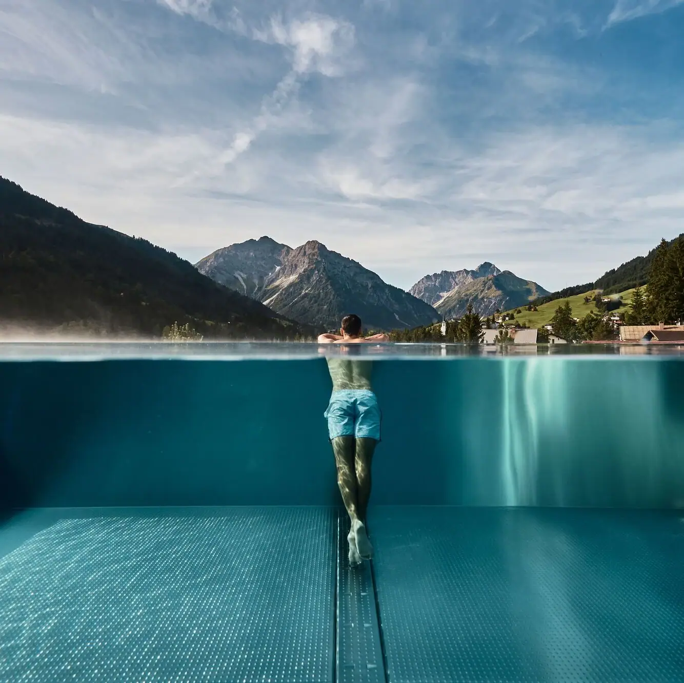 Mann lehnt am Rand eines Infinity-Pools mit Blick auf einen Ort inmitten einer grünen Berglandschaft im Hintergrund.