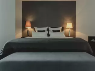 Un letto d'albergo con copriletto grigio e cuscini decorativi, piccoli comodini con lampade a luce calda e un piccolo divano in fondo al letto. 