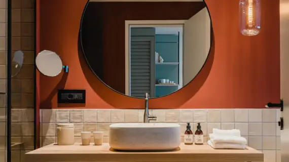 Bagno arredato con gusto e con pareti color terracotta. Al centro si trova un lavabo con un grande specchio rotondo che lo illumina. Una moderna lampada a sospensione illumina un lavabo ovale bianco su una mensola di legno chiaro. Accanto al lavabo si trovano asciugamani impilati ed eleganti prodotti per la cura della persona, che sottolineano il carattere lussuoso della stanza. Lo specchio riflette una porta aperta, offrendo uno sguardo su un'altra stanza.