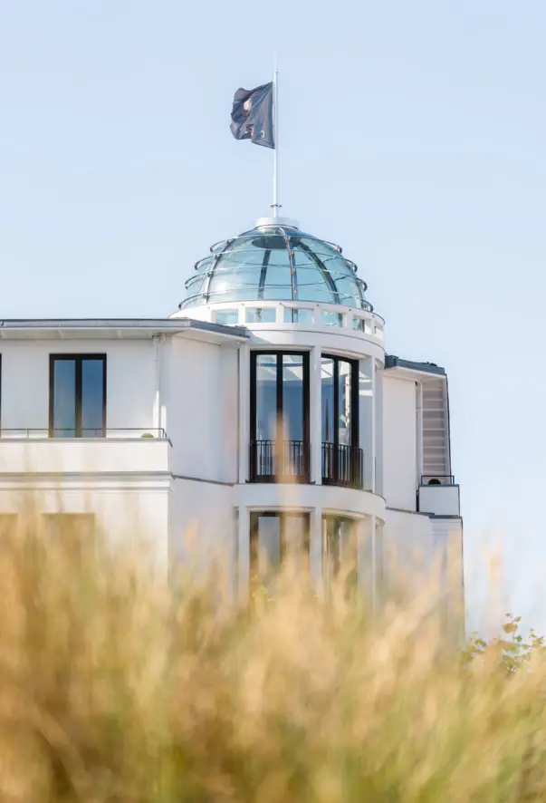 Una bandiera con la scritta "CERES" sventola al vento su una cupola di vetro di un edificio bianco. In primo piano si vedono le erbe delle dune.