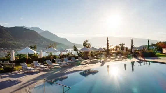 Piscina con ombrelloni e sdraio circondata da alberi e montagne con vista sul Lago di Garda.