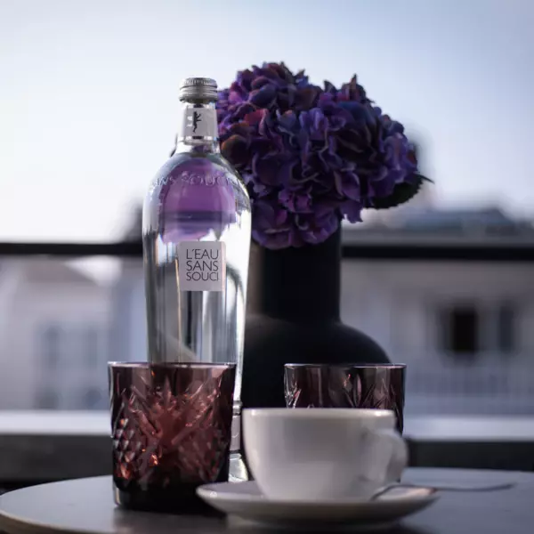 Un piccolo tavolo rotondo su un balcone, su cui sono appoggiati due tea light rosati, un vaso scuro con un rododendro viola, una bottiglia d'acqua e una tazza bianca.  