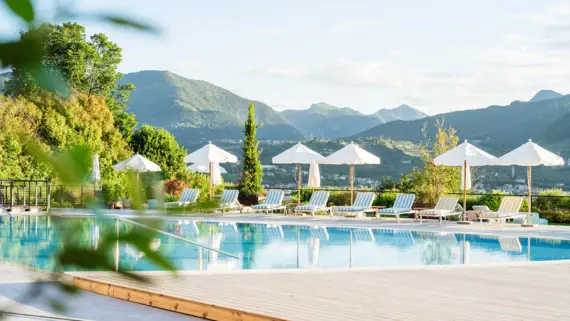 Rappresentazione fotorealistica di una lussuosa piscina all'aperto in un complesso alberghiero. Diverse persone si rilassano su sedie a sdraio e sedute di design, alcune sotto ombrelloni bianchi. Il verde lussureggiante, gli alti cipressi e gli arbusti fioriti circondano l'area della piscina, mentre un cielo azzurro con nuvole leggere adorna lo sfondo. Un edificio moderno con terrazze e tetti verdi completa l'ambiente idilliaco.