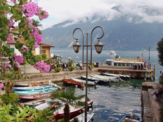 Un piccolo porto italiano con barche, una lanterna, fiori rosa e montagne sullo sfondo.