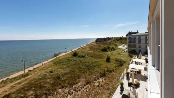 Vista sul mare, sulla passeggiata e sul paesaggio dunale. Davanti alla casa da cui è stata scattata la foto, si trova una terrazza con posti a sedere e ombrelloni.