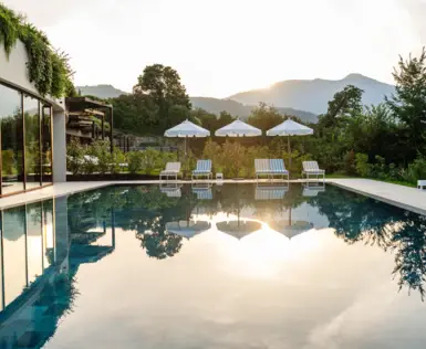 Piscina all'aperto circondata da sedie e ombrelloni con alberi e un cielo limpido con il sole al tramonto sullo sfondo.