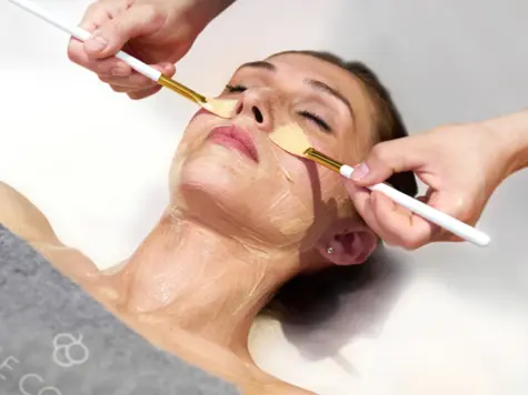 Un trattamento viso nella spa, in cui le mani di un'estetista applicano con un pennello una maschera nutriente sul viso di una cliente rilassata. La cliente, con gli occhi chiusi, si gode visibilmente il trattamento. L'immagine trasmette una sensazione di lusso, relax e cura professionale della pelle.