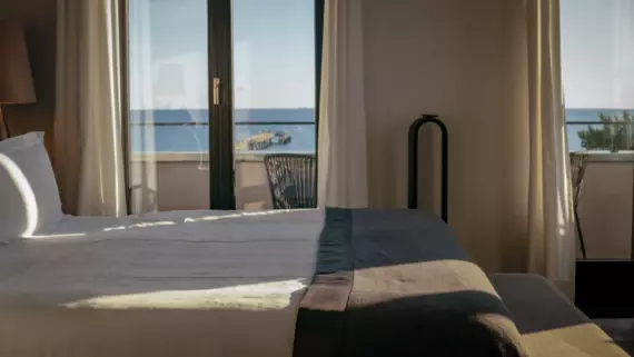 Una camera d'albergo esposta al sole con finestre a tutta altezza. Dal lato si vede un grande letto bianco con un copriletto scuro e il mare aperto sullo sfondo. 