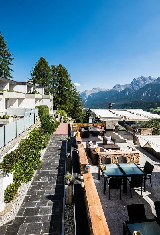 Patio esterno con tavoli e sedie, circondato da alberi e con montagne sullo sfondo.