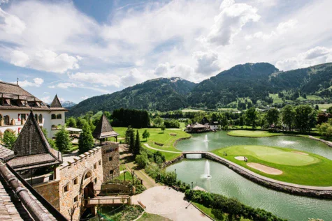 Campo da golf con fontana e un edificio sullo sfondo, circondato da alberi e montagne.