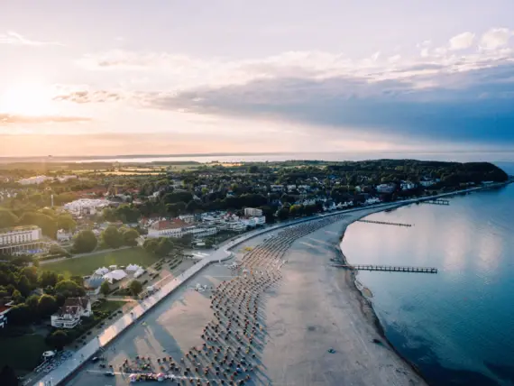 Una veduta aerea della zona costiera di Travemünde. L'acqua è di un caldo colore blu, la spiaggia si estende lungo la costa e molte sedie a sdraio sono sparse lungo di essa, mentre la città giace tranquillamente sullo sfondo tra il verde. 