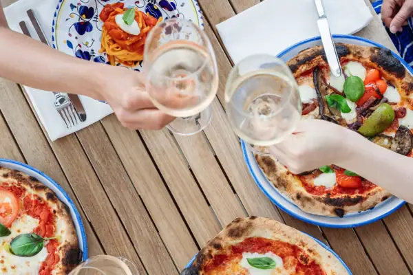 Hände stoßen mit Weingläsern an, daneben frisch gebackene Pizza auf einem Holztisch.