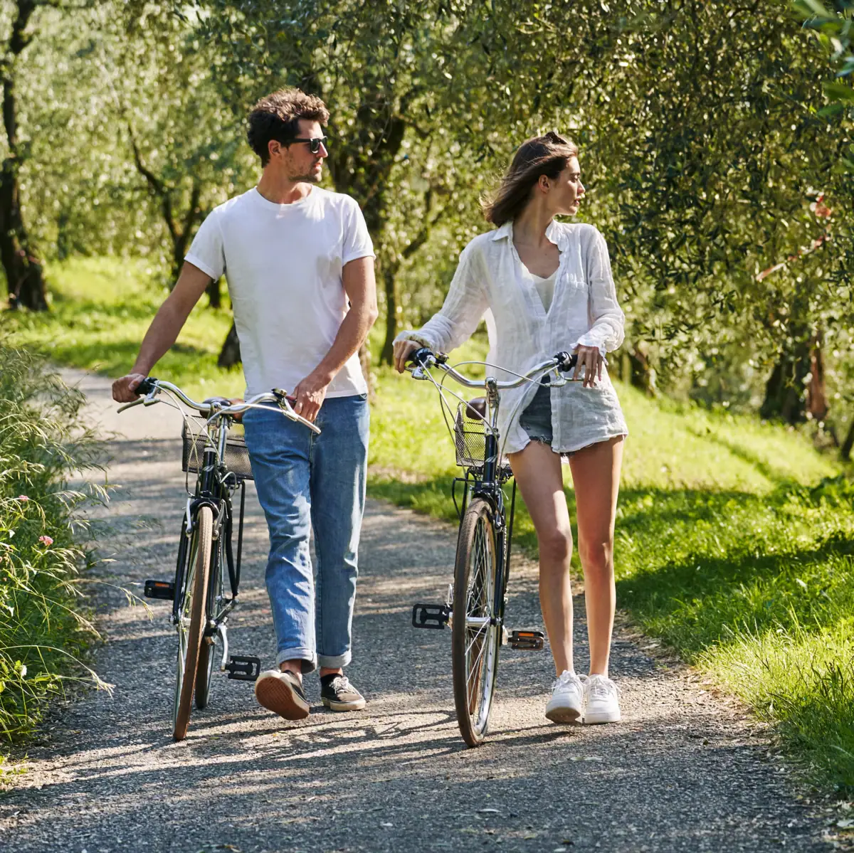 Una coppia passeggia con le biciclette attraverso un uliveto soleggiato vicino al Lago di Garda – natura, relax e vacanza attiva nella regione della Valtenesi.