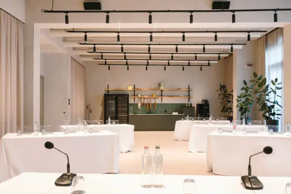 Ein Tagungsraum mit Tischen und Stühlen. Der vordere Tisch ist mit Mikrofonen sowie Gläsern und Wasserflaschen eingedeckt. Die hinteren Tische sind auf mit Gläsern und Wasserflaschen eingedeckt. Ganz hinten im Raum sieht man einen Kühlschrank und eine Küchenzeile für eine Kaffeepause.