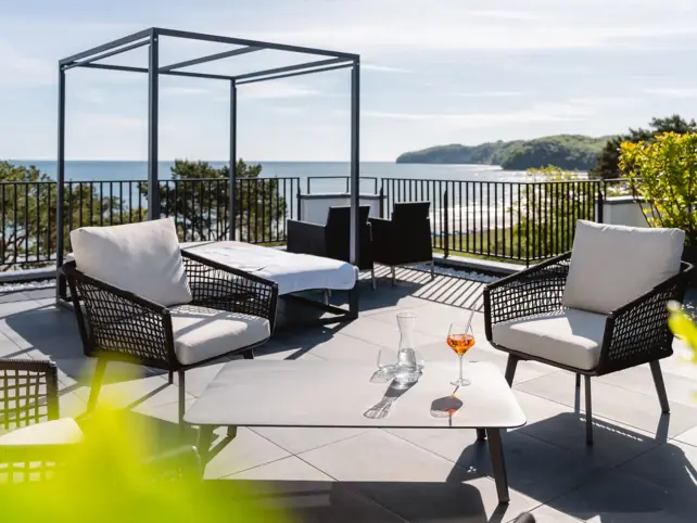 Una terrazza sul tetto di un edificio bianco con finestre nere in stile francese. Alcuni ombrelloni bianchi sono stesi sulla terrazza verde del tetto e si vedono alcuni lettini. Sullo sfondo si vedono il Mar Baltico e il cielo al tramonto.