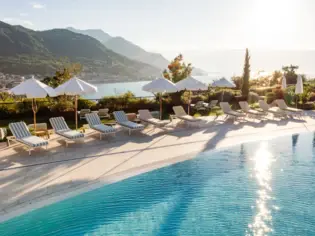 Piscina con sedie e ombrelloni in riva al mare, circondata da montagne e alberi.