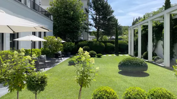 Un giardino ben curato con un prato immacolato, fiancheggiato da cespugli sferici accuratamente tagliati e da alcuni giovani alberi, davanti a un elegante edificio. Davanti all'edificio sono stesi degli ombrelloni bianchi. 