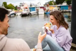 Una donna siede su un molo e mangia un hamburger.