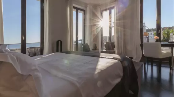 Una camera da letto soleggiata con molte grandi finestre. Al centro della stanza si trova un grande letto rivestito di bianco con vista sul mare.  