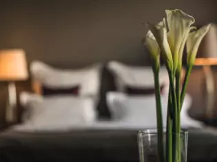 In primo piano un vaso di eleganti calle bianche. Sullo sfondo sfocato si intravede una camera da letto, illuminata dalle luci soffuse delle lampade da tavolo ai lati di un letto matrimoniale con lenzuola bianche e cuscini neri.