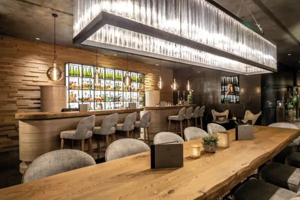 Ein langer Holztisch mit Stühlen vor einem Bartresen mit Barhockern vor einer gut gefüllten Bar im Hintergrund..