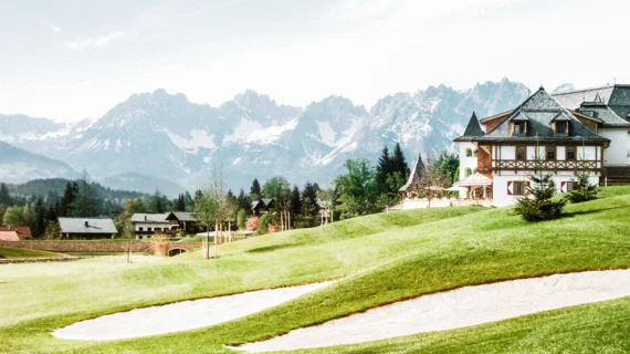 Campo da golf con edificio e montagne sullo sfondo, circondato da alberi e cielo nuvoloso.