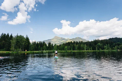 Persona su una tavola da paddle in un lago circondata da alberi e montagne.