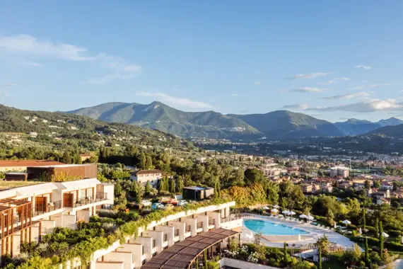 Una veduta della pittoresca cittadina di Salò sul Lago di Garda, circondata da dolci colline e da una fitta vegetazione. L'architettura degli edifici sulla riva trasmette un'atmosfera classica italiana, mentre le montagne sullo sfondo evocano un senso di avventura e di vicinanza alla natura. 