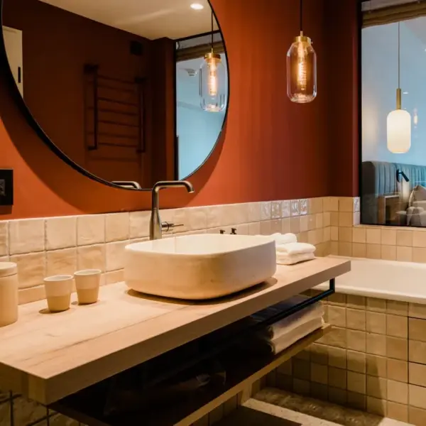 Un bagno elegante con parete color terracotta e zona lavaggio piastrellata in beige. Un grande specchio rotondo è appeso sopra il lavabo con vasca ovale in pietra e sotto c'è una mensola in legno aperta con gli asciugamani. Due lampade a sospensione illuminano l'ambiente, mentre la camera da letto con letto matrimoniale è visibile attraverso una parete di vetro sullo sfondo.