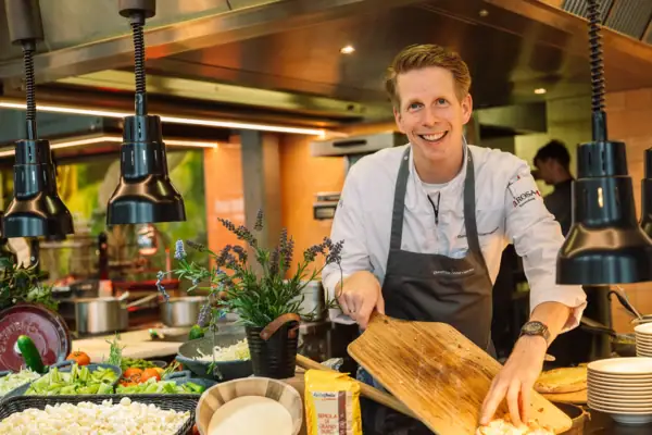 Ein Koch mit Kochschürze steht in einer Küche, hinter dem Buffet und hält ein Schneidebrett in der Hand.