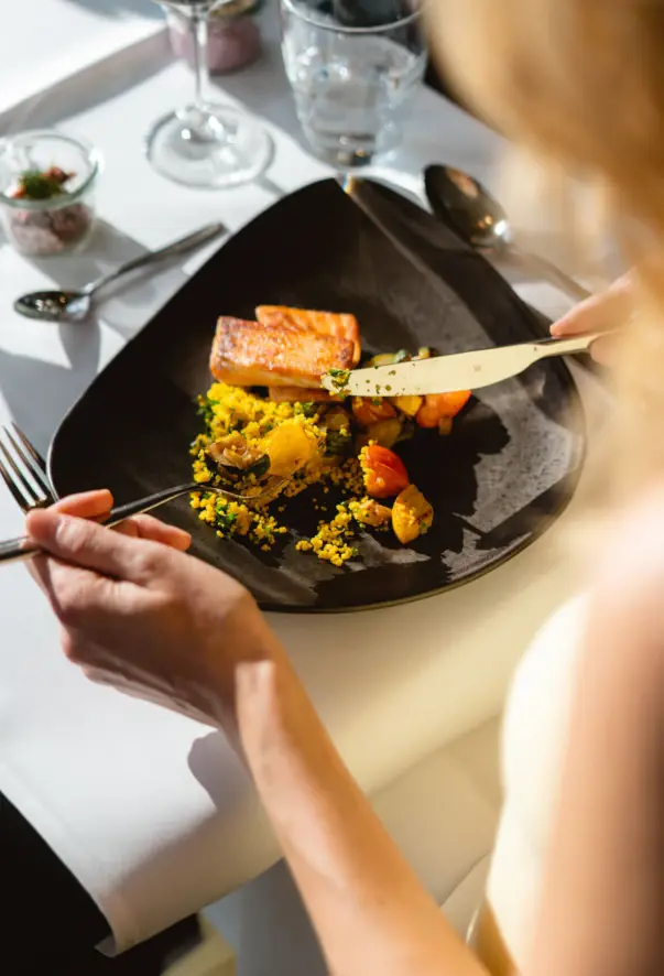 Persona che mangia un pasto su un piatto con posate, bicchiere di vino e altri elementi da tavola.