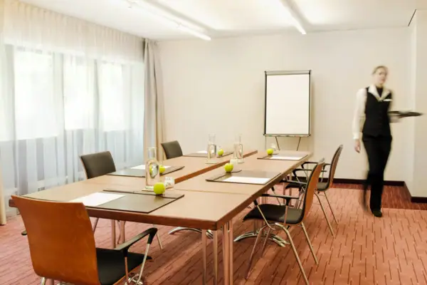 Eine Frau steht in einem Tagungsraum mit einem Tischblock und Stühlen drumherum.