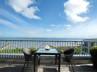 Una terrazza con un tavolo e due sedie davanti alla ringhiera. Sullo sfondo si vedono il mare e il cielo azzurro.