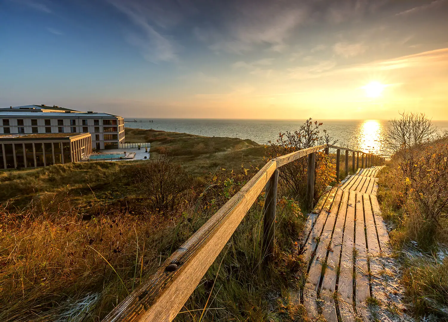 Holzsteg führt zu einem Strand mit einem Gebäude und einem Gewässer im Hintergrund.