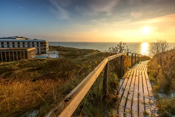 Holzsteg führt zu einem Strand mit einem Gebäude und einem Gewässer im Hintergrund.