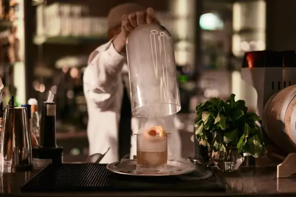 Person nimmt eine Glashaube über einem dampfenden Glas mit einem Drink hoch, umgeben von einer Bar mit einer Pflanze auf dem Tresen in einem Innenraum.