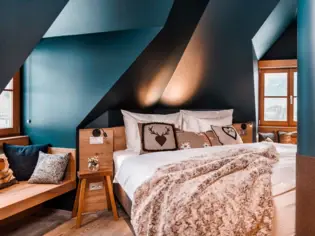 Un letto accogliente con una coperta marrone e tre cuscini decorativi in stile alpino. A sinistra del letto si trova un piccolo comodino in legno e una zona salotto con cuscini.