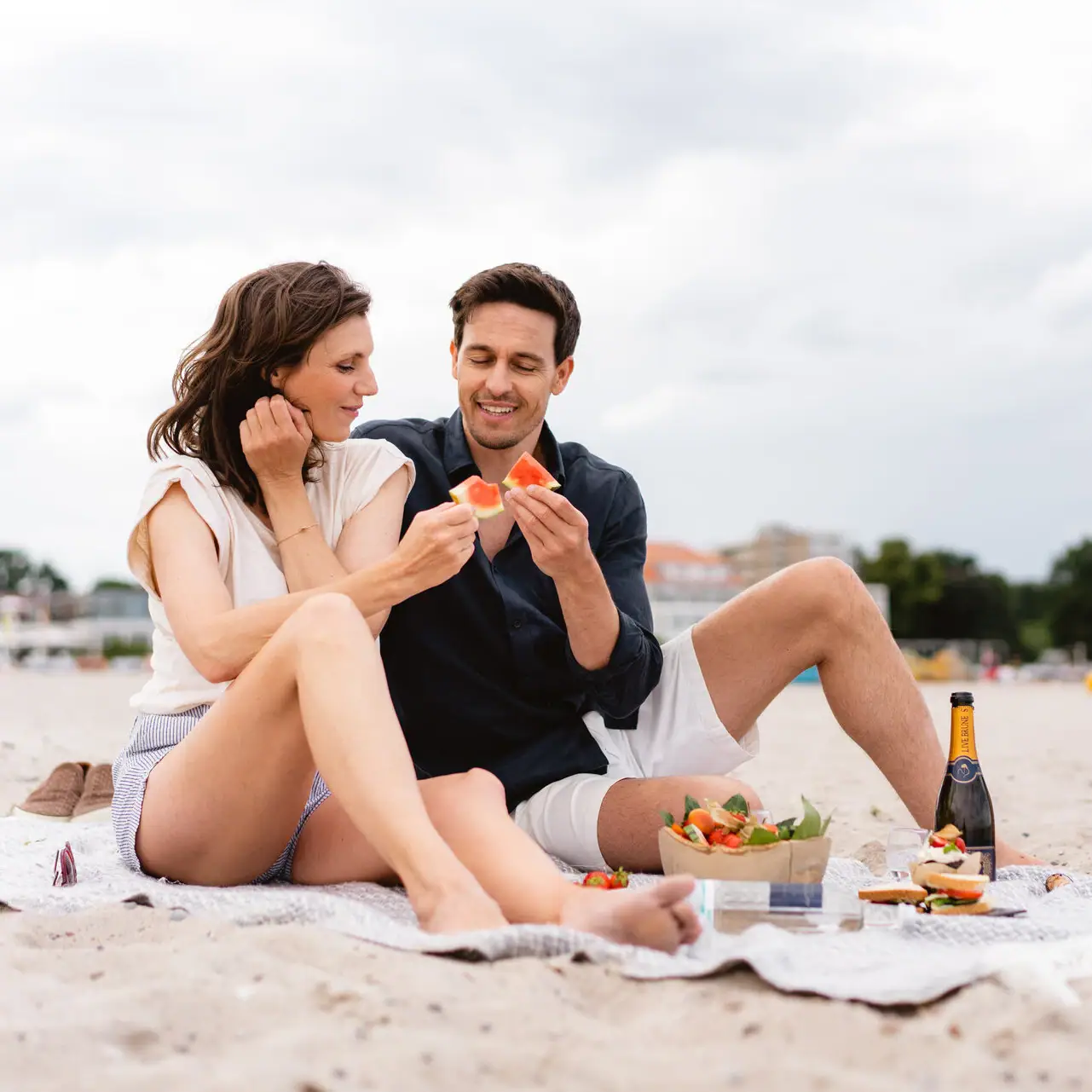 Un uomo e una donna seduti su una coperta in spiaggia mentre mangiano.